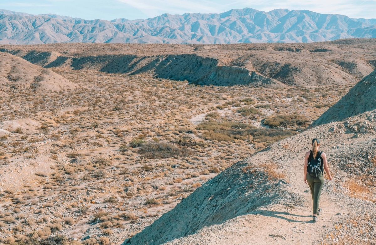 A woman wearing a backpack and green pants walking away though a mountainous, desert landscape.