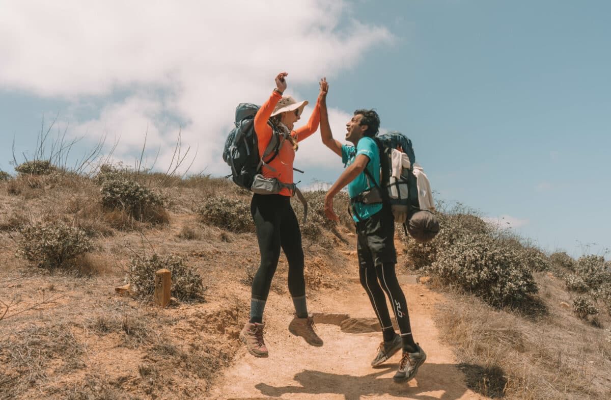 A woman wearing an orange long-sleeved hiking shirt, black leggings, and a backpack jumps in the air to high-five a man in an aqua shirt with a dusty trail and a blue sky behind them.