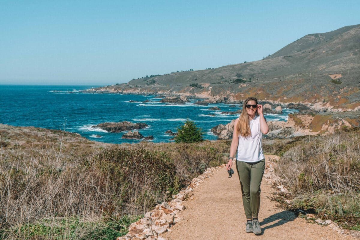 A woman wearing, sunglasses, dark green pants, and a white tank smiles as she walks along an oceanside path with a beach and hills in the background.