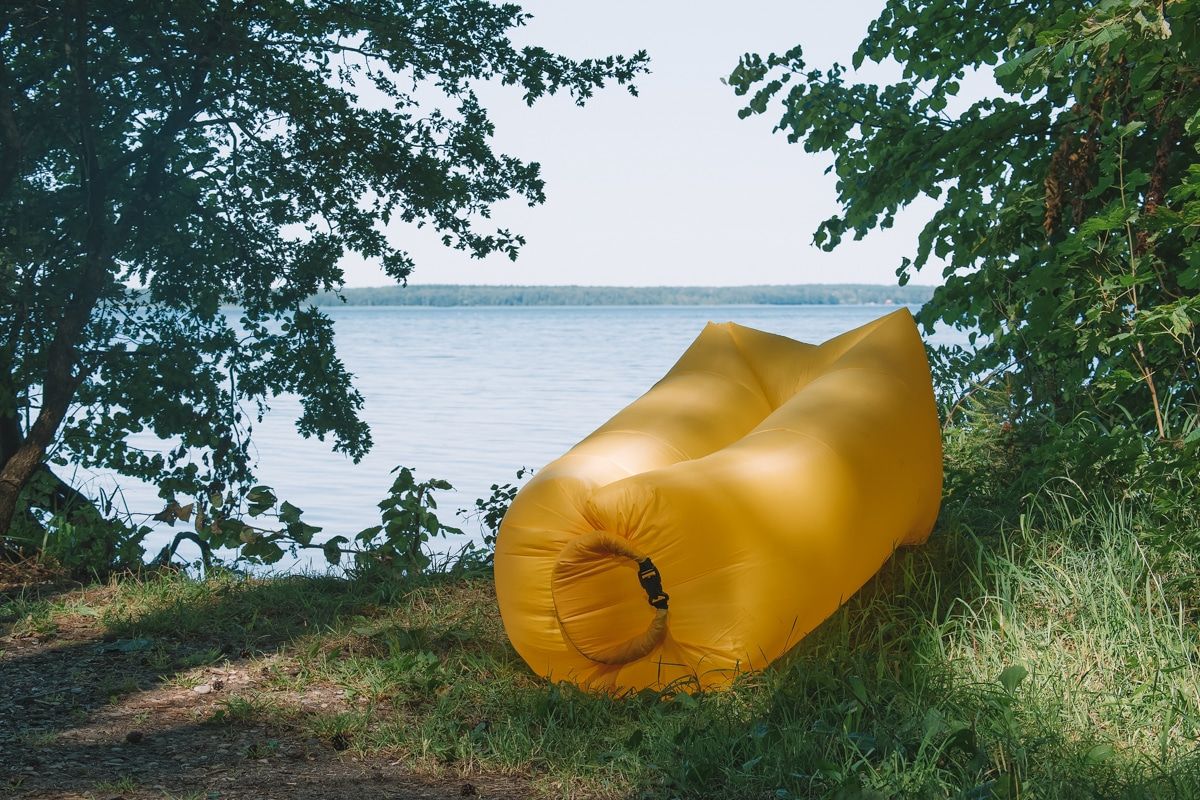 A yellow inflatable lounger sits on grass surround by leafy branches overlooking a lake.