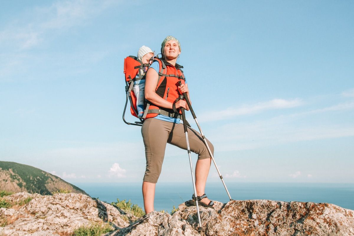 A woman wearing cropped leggings and an orange vest carries a baby in an orange backpack carrier and steps over rocky terrain with an ocean view behind her.