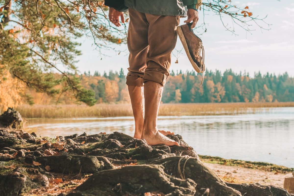 The lower half of a man wearing rolled up khaki pants, holding a pair of sneakers, and standing barefoot on some tree roots on the bank of a lake, with autumn leaves in the background.