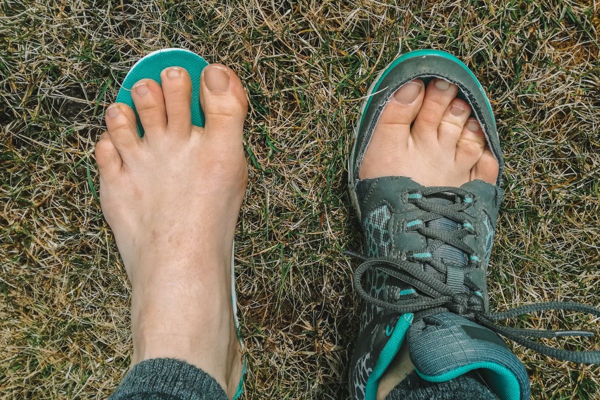 A pair of feet standing on a lawn, one bare and one inside a sneaker with the top cut away to show how conventional shoes constrict the feet.