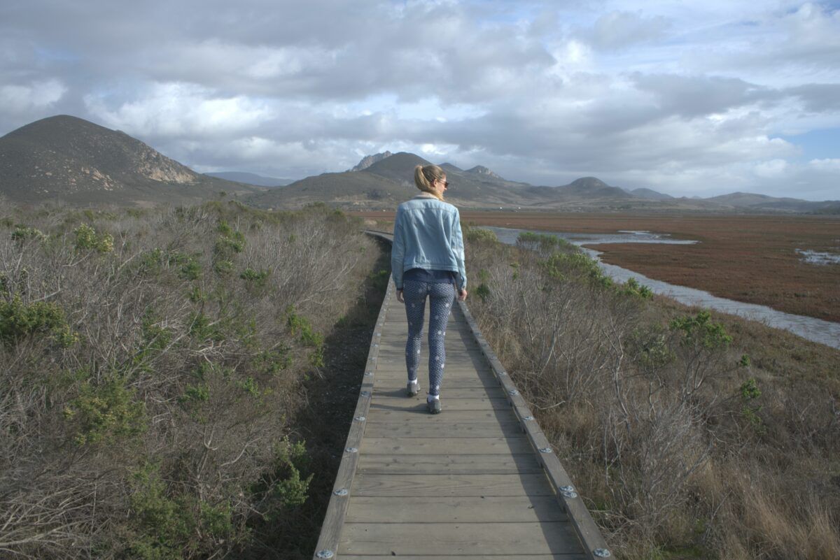 A woman wearing patterned grey leggings and a jean jacket seen from behind walking along a wooden boardwalk path through a marshy landscape with rolling hills in the distance.