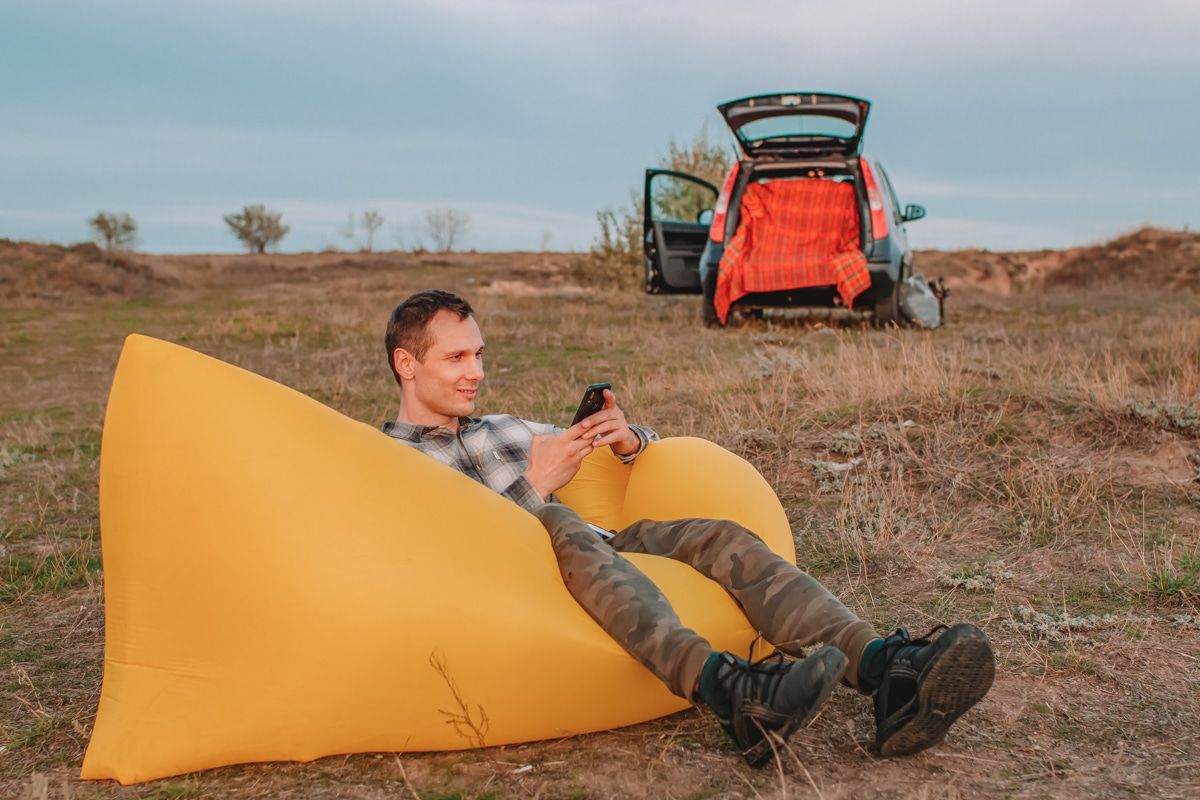 A man looks at his phone while sitting on a yellow inflatable lounger in a campsite, with an SUV with an open hatchback in th ebackground.