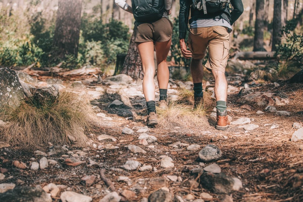 The lower halves of a male hiker and a female hiker, both wearing brown khaki shorts walking along a rocky trail through the forest.