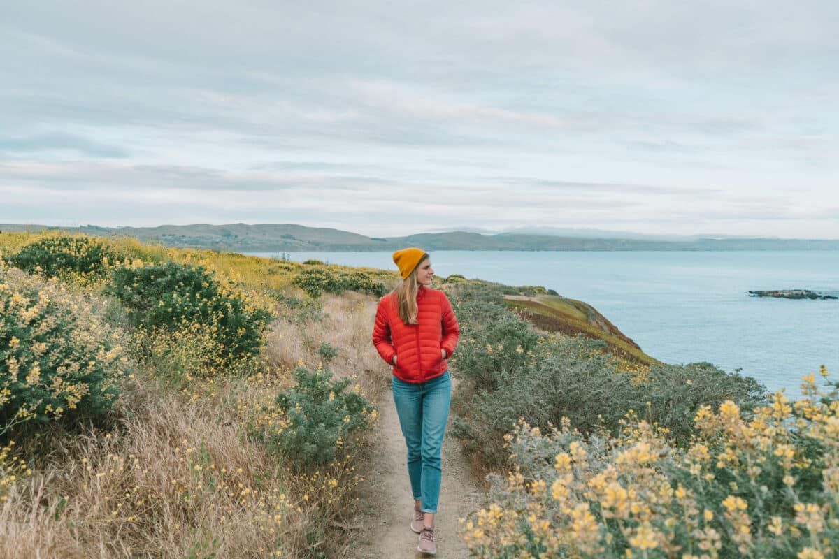 A woman in jeans, a red puffy jacket, and a yellow beanie walks along a trail lined by bushes overlooking the ocean beneath an overcast sky.