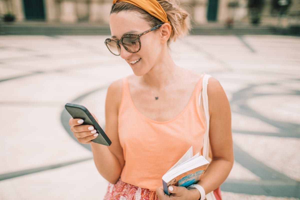 A young woman in a peach tank top and sunglasses holds a travel guide book under one arm, and checks her phone.