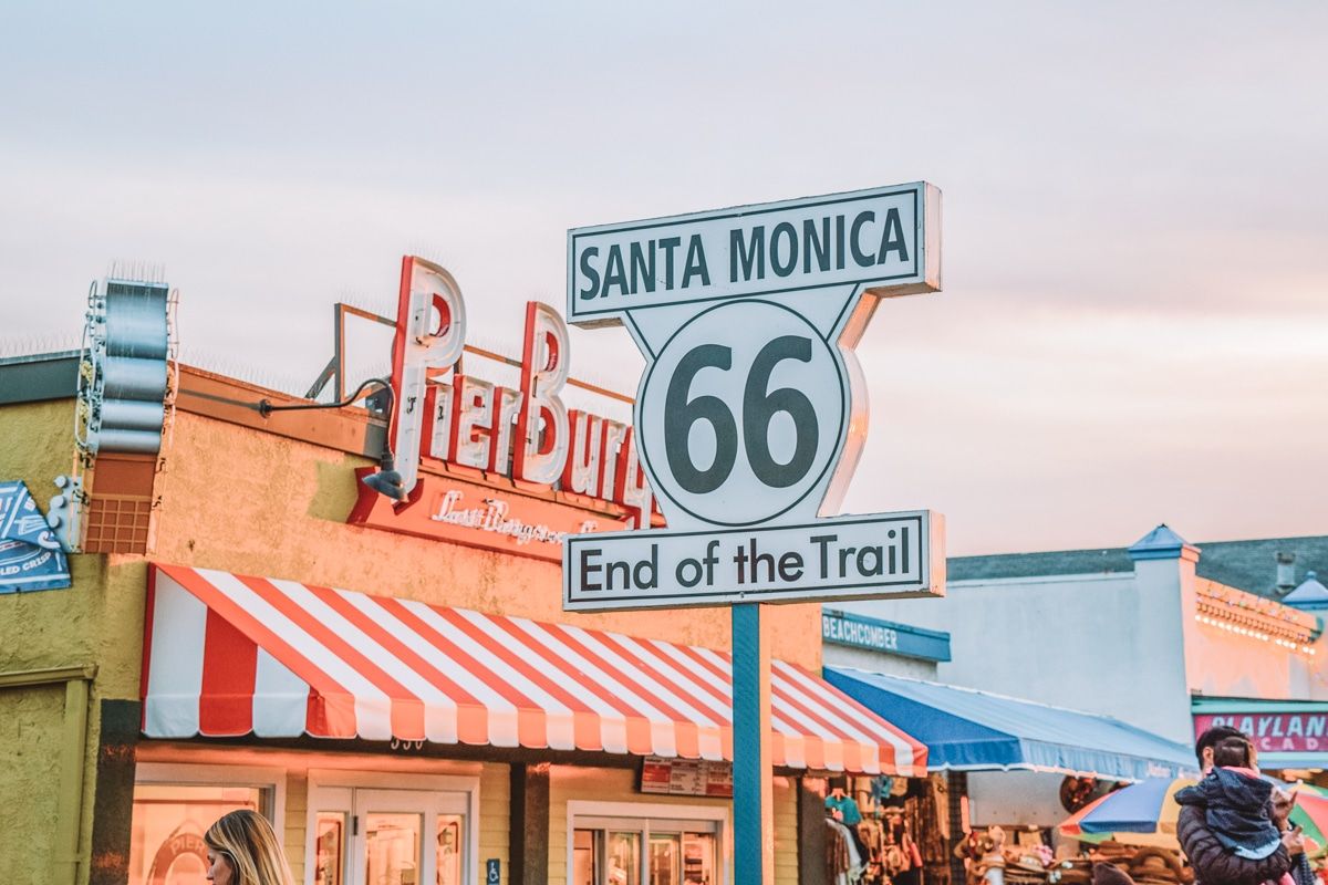 The black-and-white Route 66 Street Sign with the red-and-white striped awning of Pier Burger in the background.