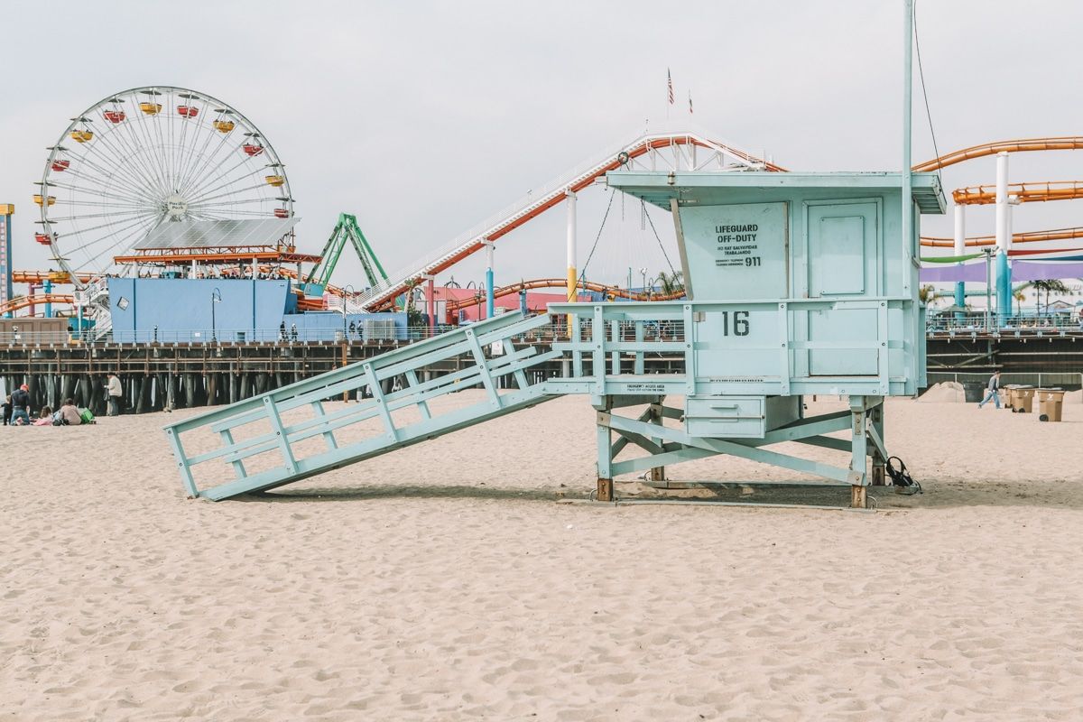 A light blue lifeguard's office on the beach, with the rides at the Santa Monica Pier boardwalk behind it.