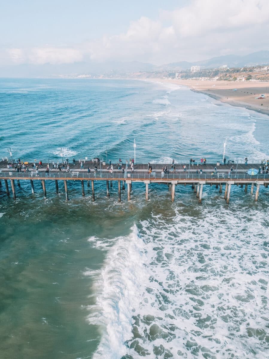 A bird's eye view of people walking on the dock over blue-green waves at the Santa Monica Pier.