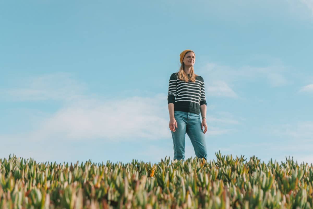 A woman in jeans, a yellow beanie,  and a striped shirt stands amongst ice plants on a hilltop with a blue sky behind her.