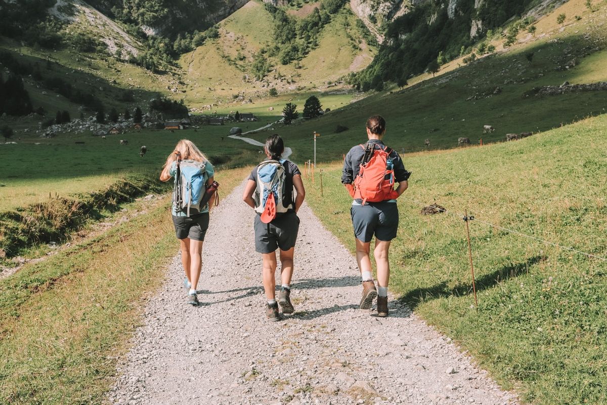 Three female hikers wearing shorts and backpacks seen from behind walking abreast on a wide, pebbly trail leading through a grassy valley.