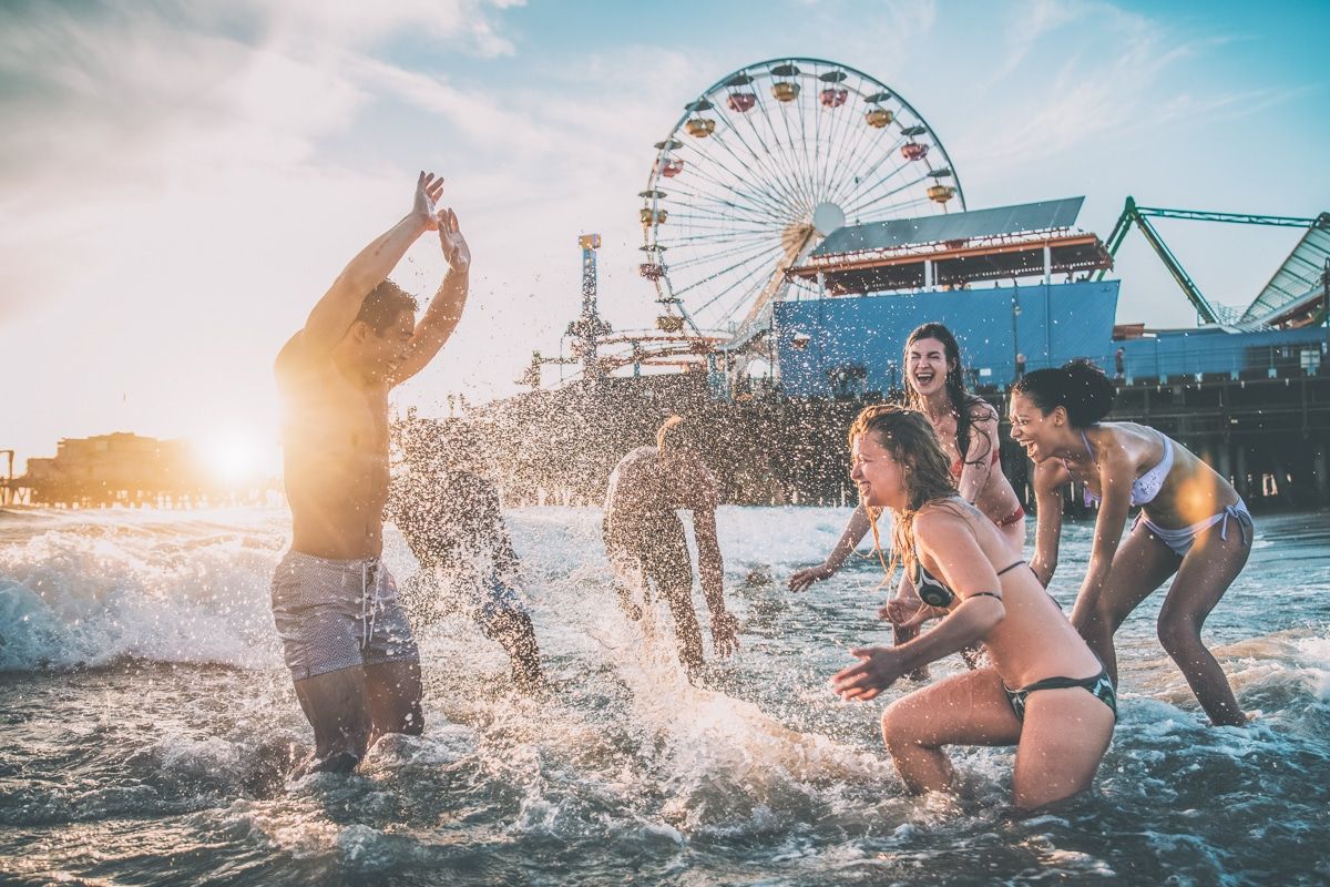 Six young people in bathing suits laughing and splashing water at the each other in the waves at the beach, with the Santa Monica Pier behind them.