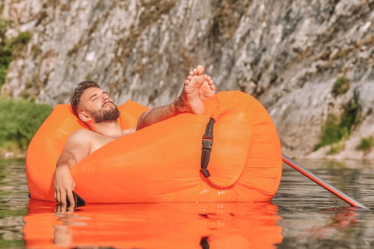A man relaxing on an orange inflatable lounger that's floating on a lake, with a rocky cliffside in the background.