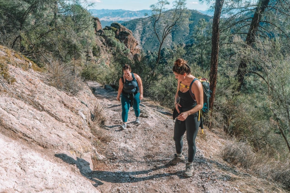 Two women wearing leggings and backpacks hiking up a rocky, tree-lined trail in the mountains.