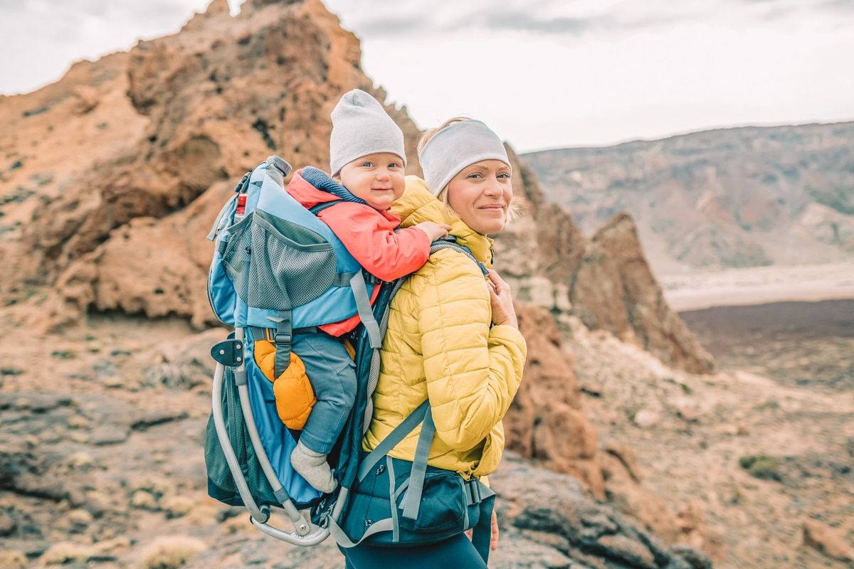 A blonde woman wearing a yellow down jacket carries a baby in a backpack-style carrier in a rocky landscape.