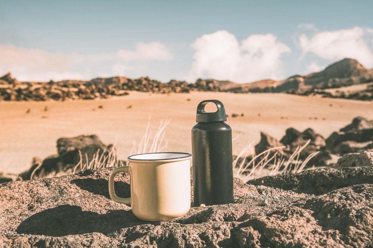 A white enamel camping mug with a black stainless steel water bottle sitting beind it on some rocks in a desert landscape.