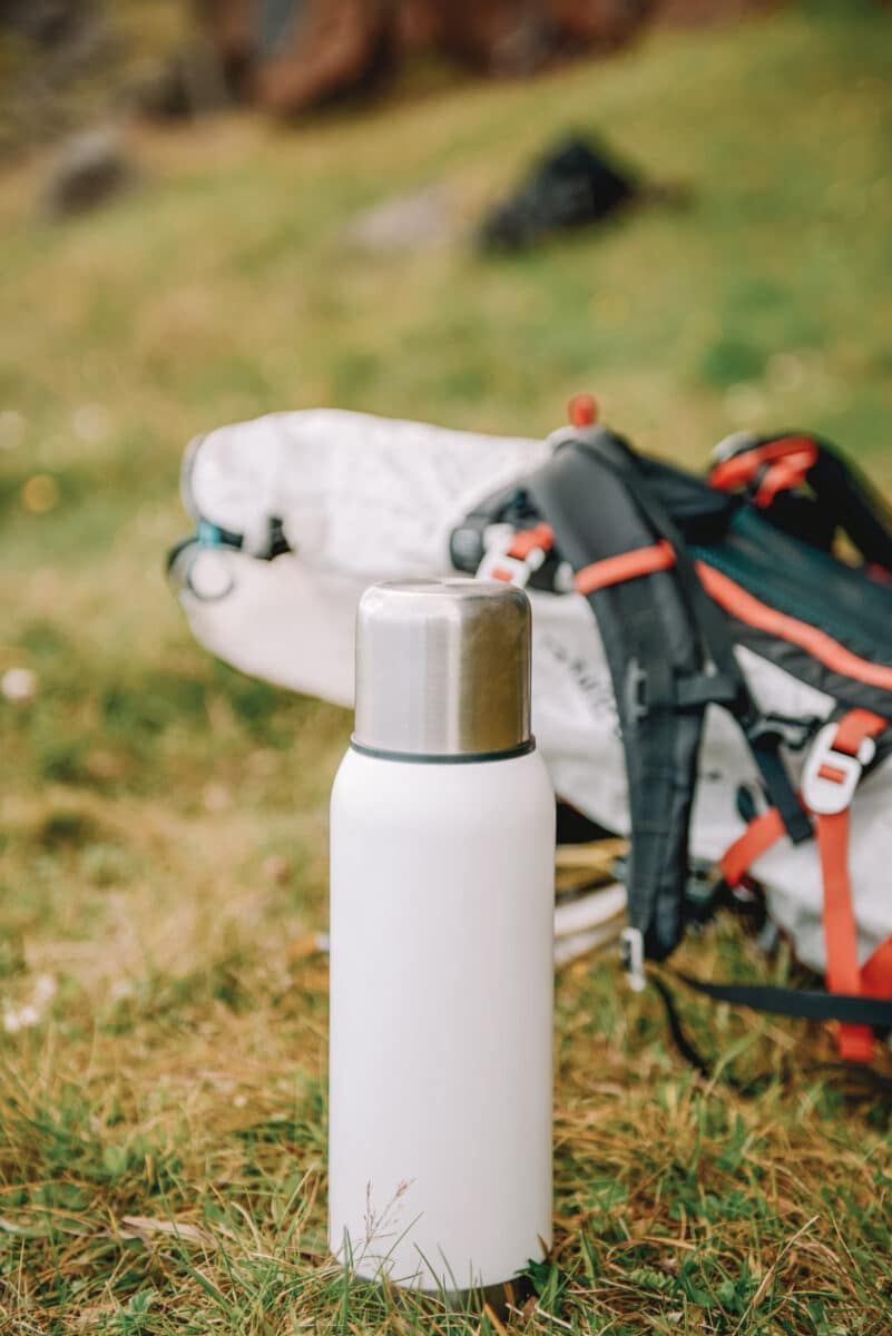 An example of one of the the best hydro flask alternative water bottles, a white stainless steel bottle, sitting on grass with a hiking backpack behind it.