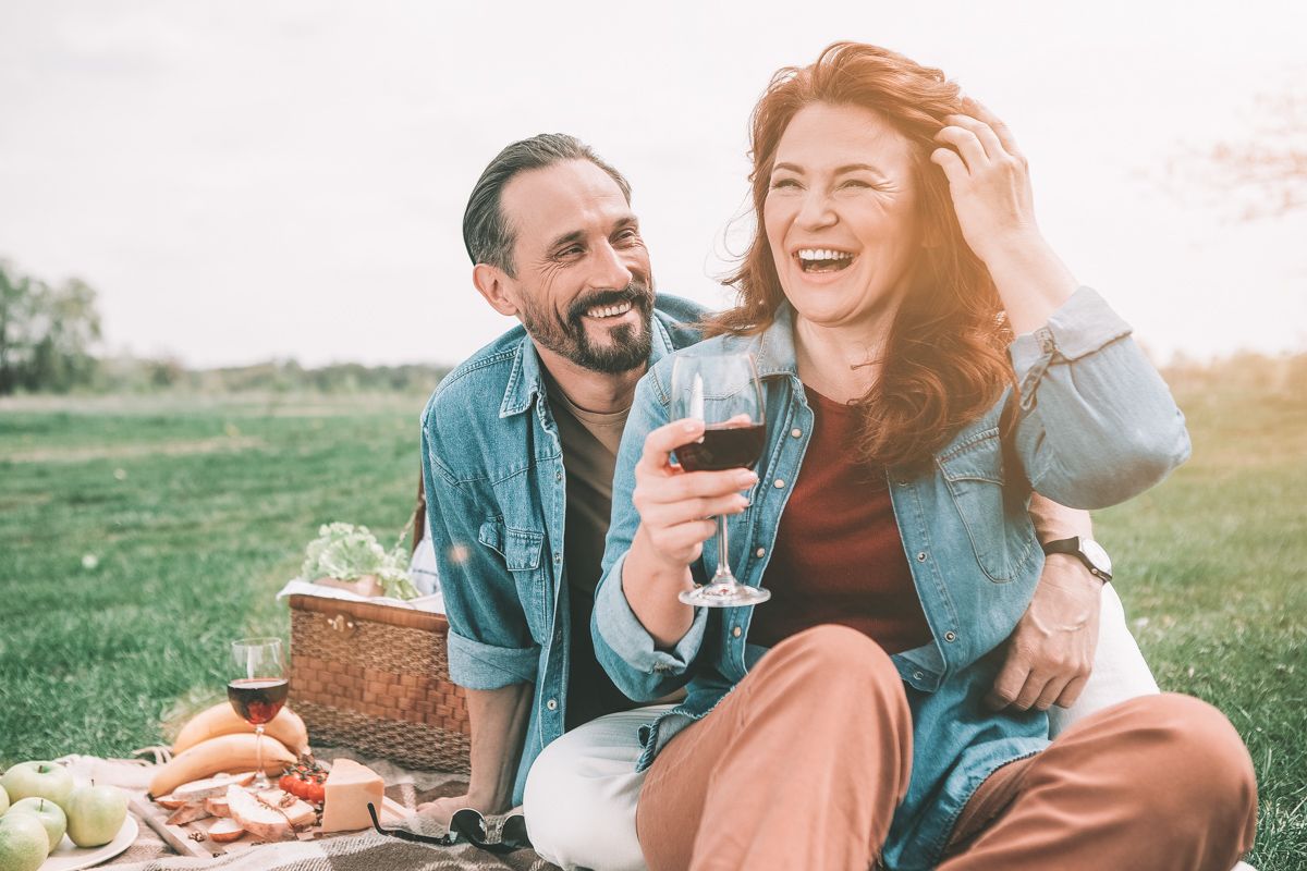 A middle-aged couple, both wearing denim shirts, laughs while enjoying red wine and a picnic in a grassy field.