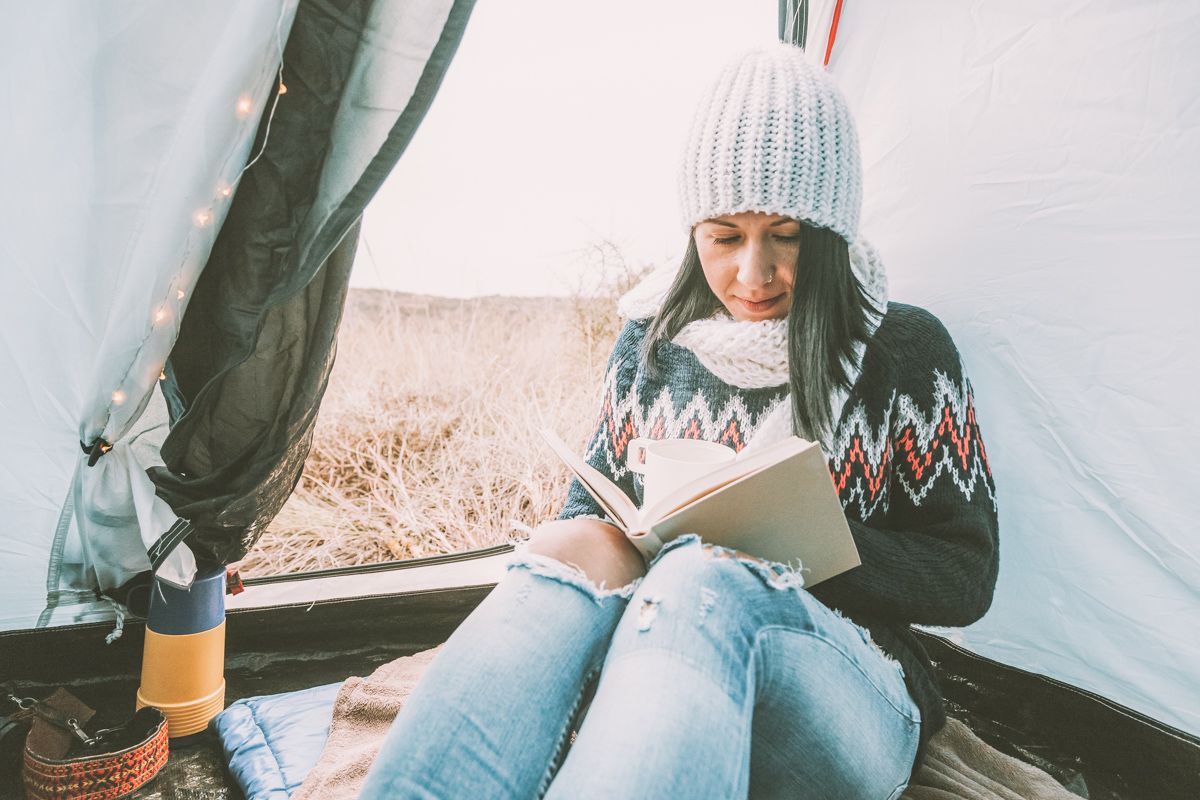 A dark-haired young woman bundled in a sweater, scarf, and cap reading a book in the doorway of a tent with a rainy-looking landscape visible outside.