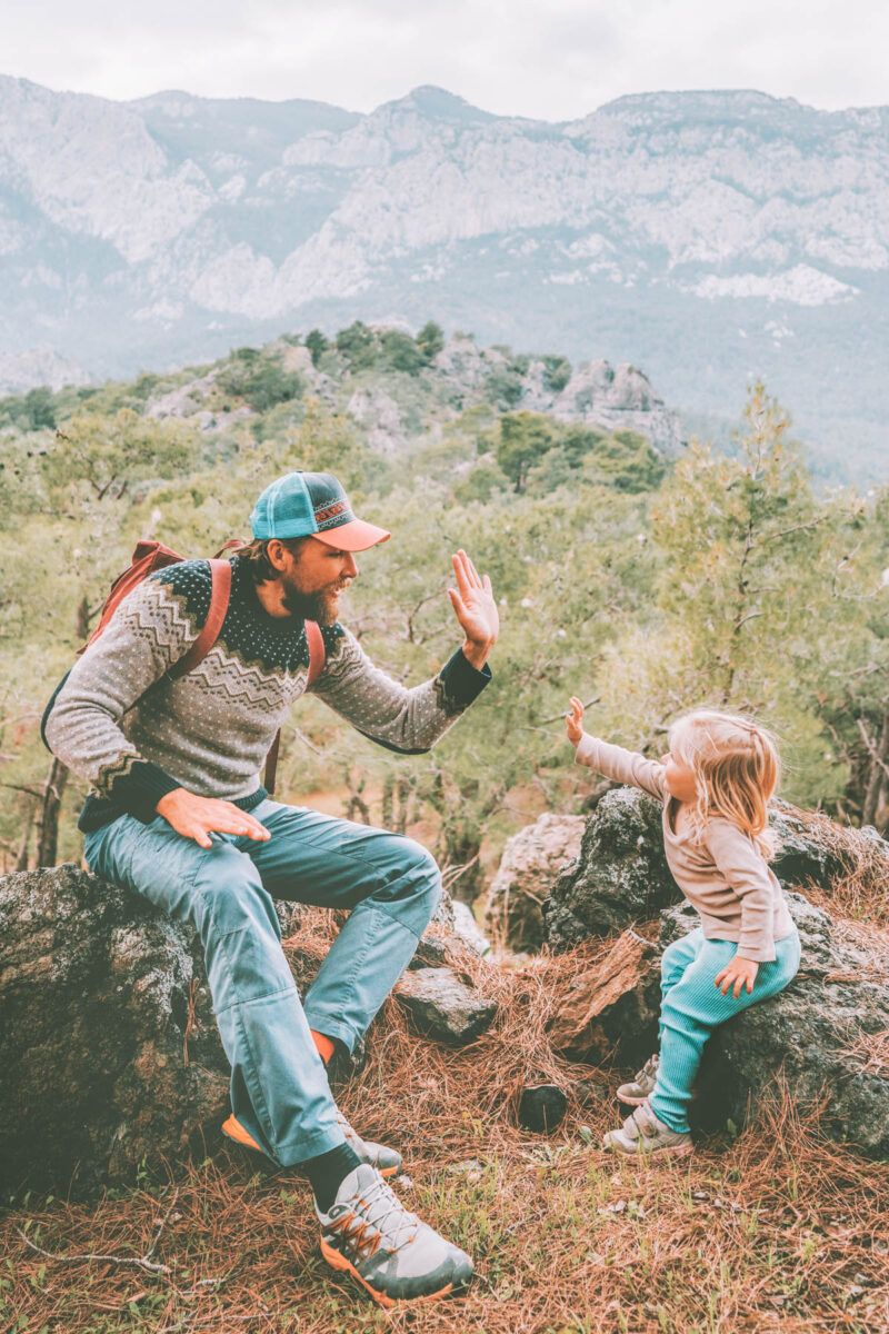 Best Camping Activities: A man in a patterned sweater and a blue and orange baseball cap high-fives a blond toddler as they sit on rocks covered in pine-needles overlooking a forested mountain vista.