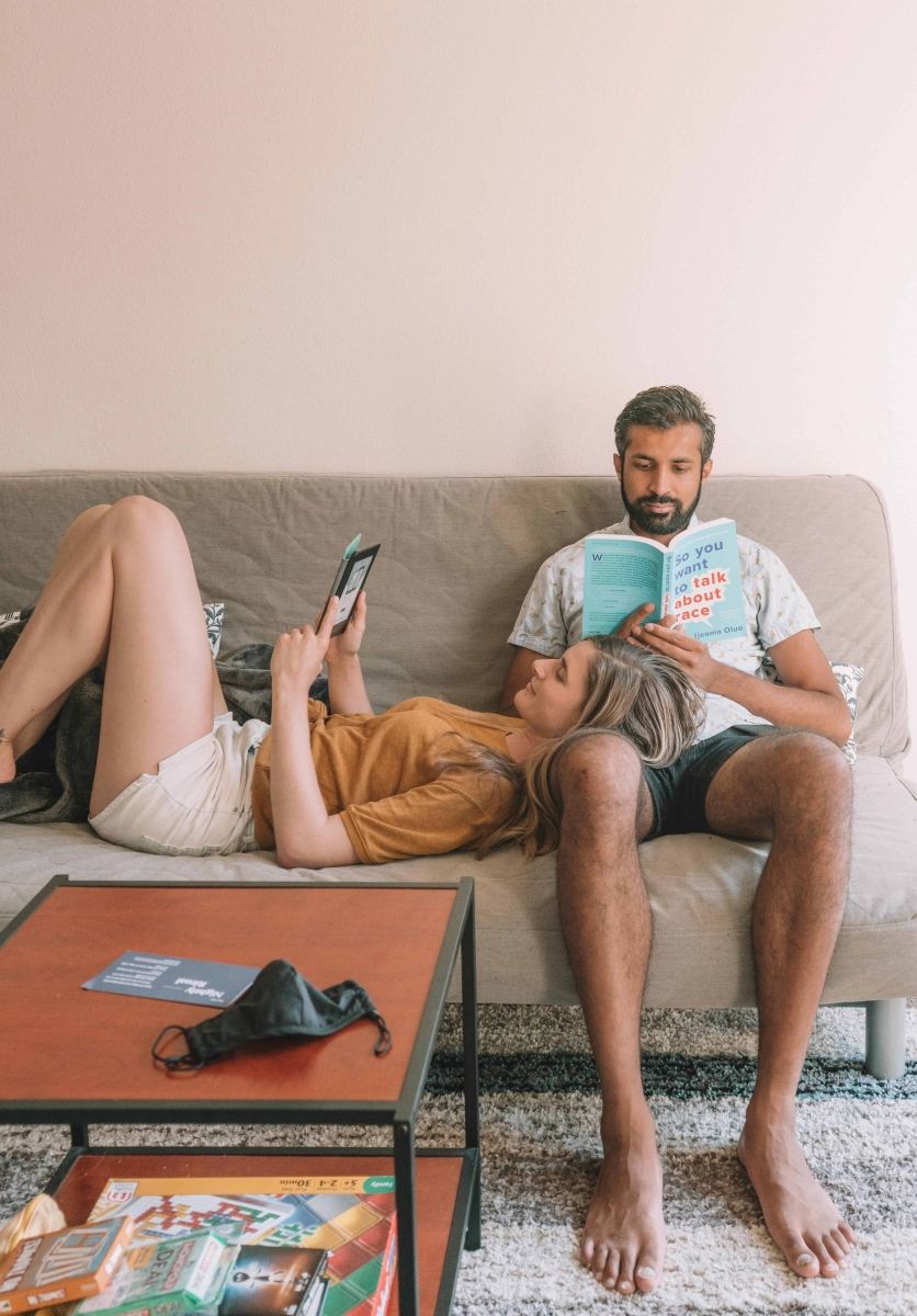 A couple reading on the couch. The woman is laying down with her head on the man's leg as she reads an e-book. The man sits upright reading a book titled "So You Want To Talk About Race."