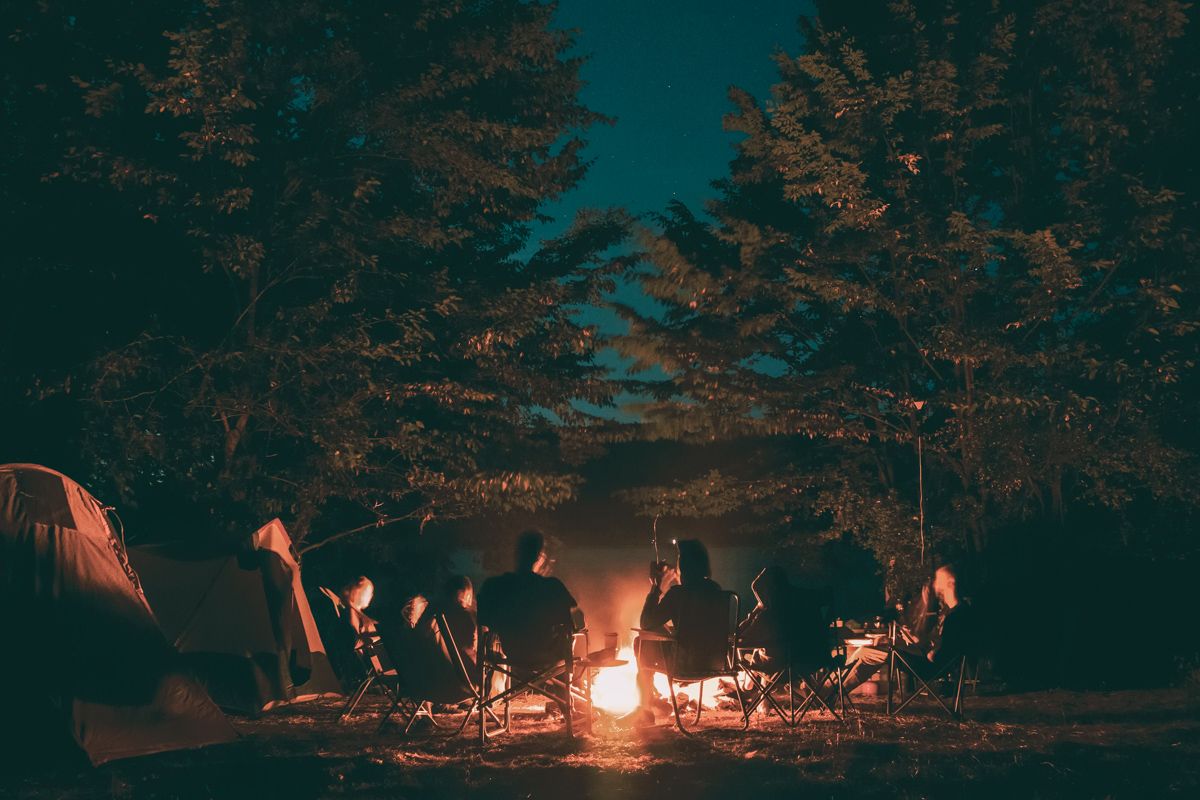A group of people sit on folding camping chairs surrounding a campfire with trees and the night sky above them.