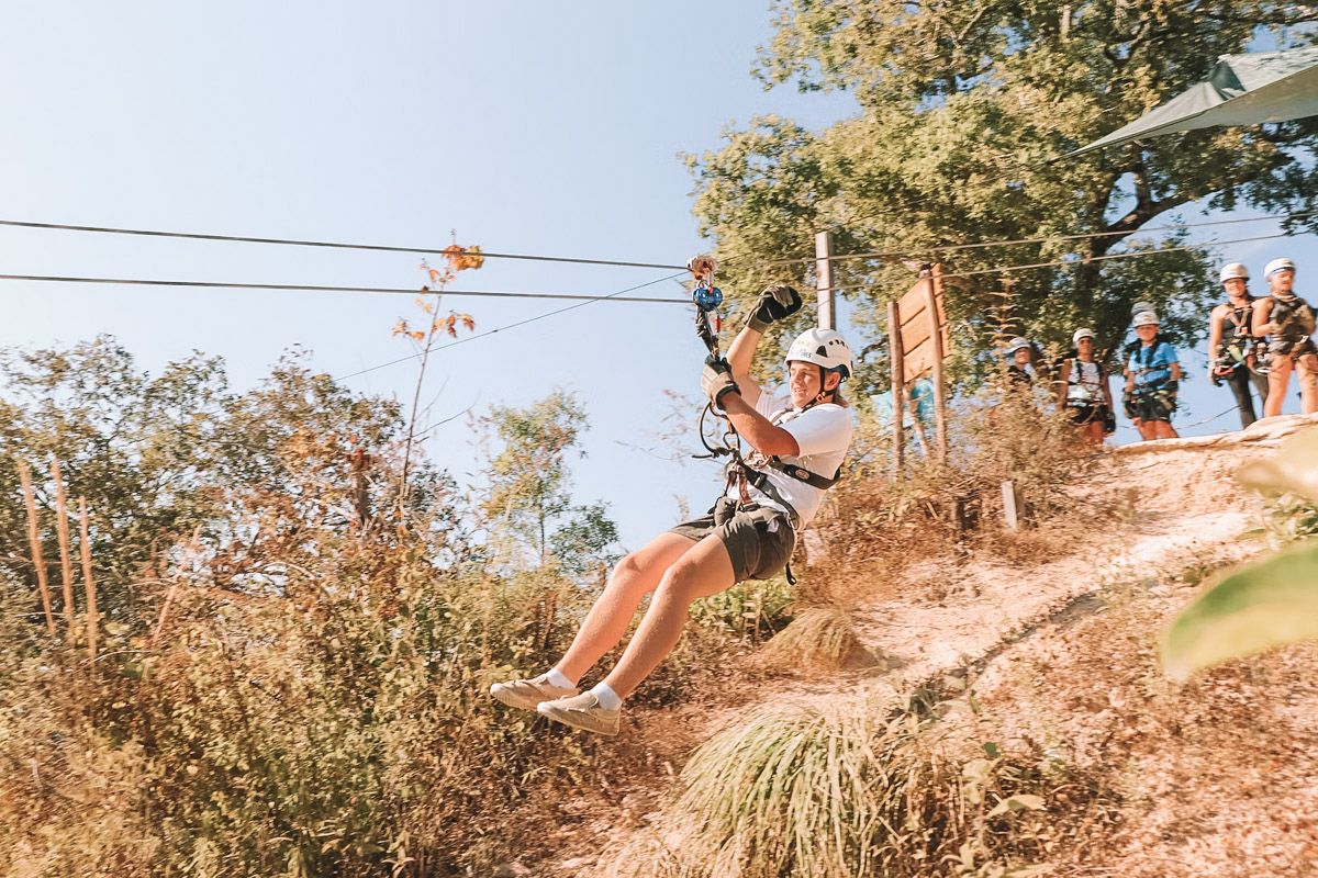 A person wearing shorts and a while t-shirt rides a zipline over dry California vegetation with a clear blue sky behind them.