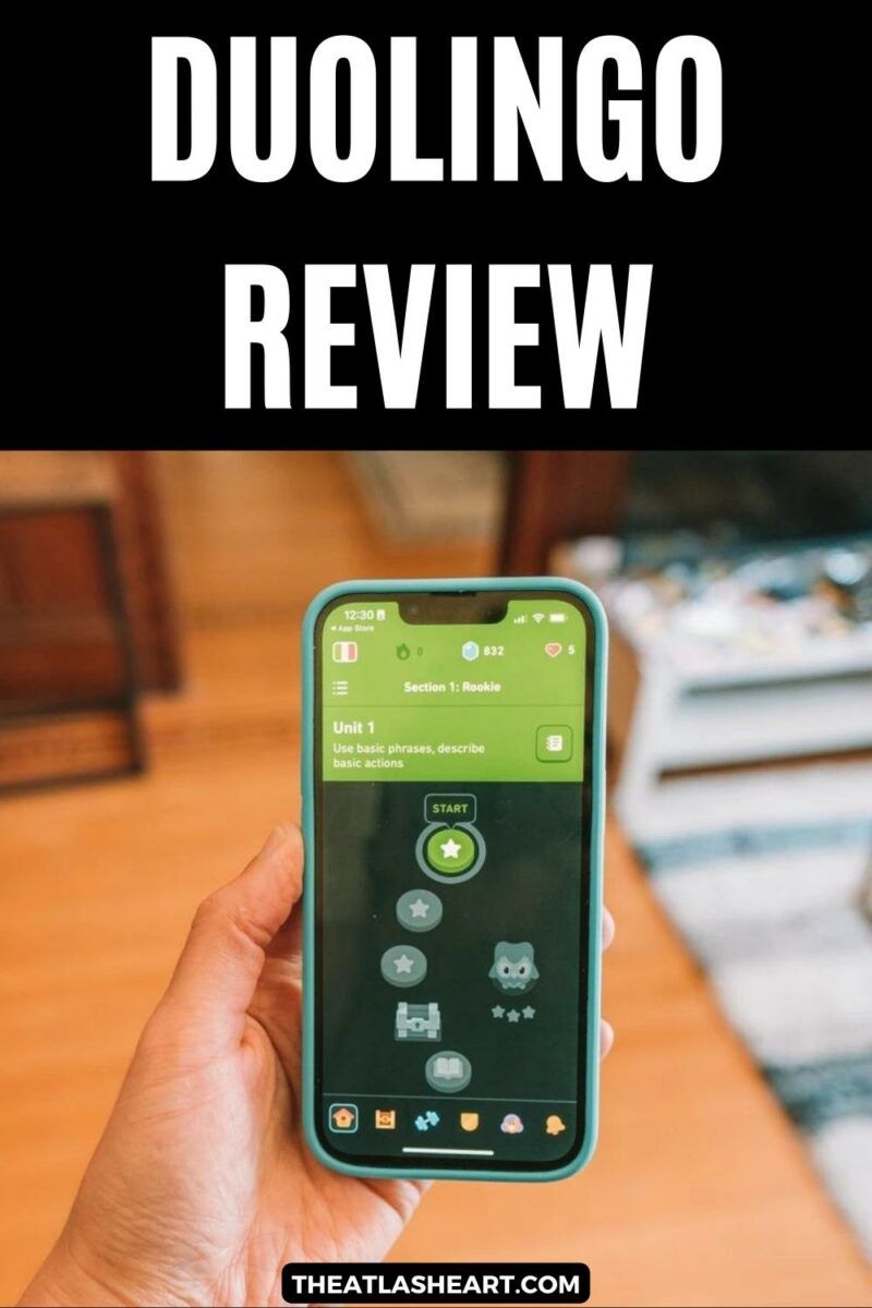 A hand holding an iPhone displaying Section 1: Rookie page in the Duolingo app, and a soft-focus apartment interior visible in the background, with the text overlay, "Duolingo Review."