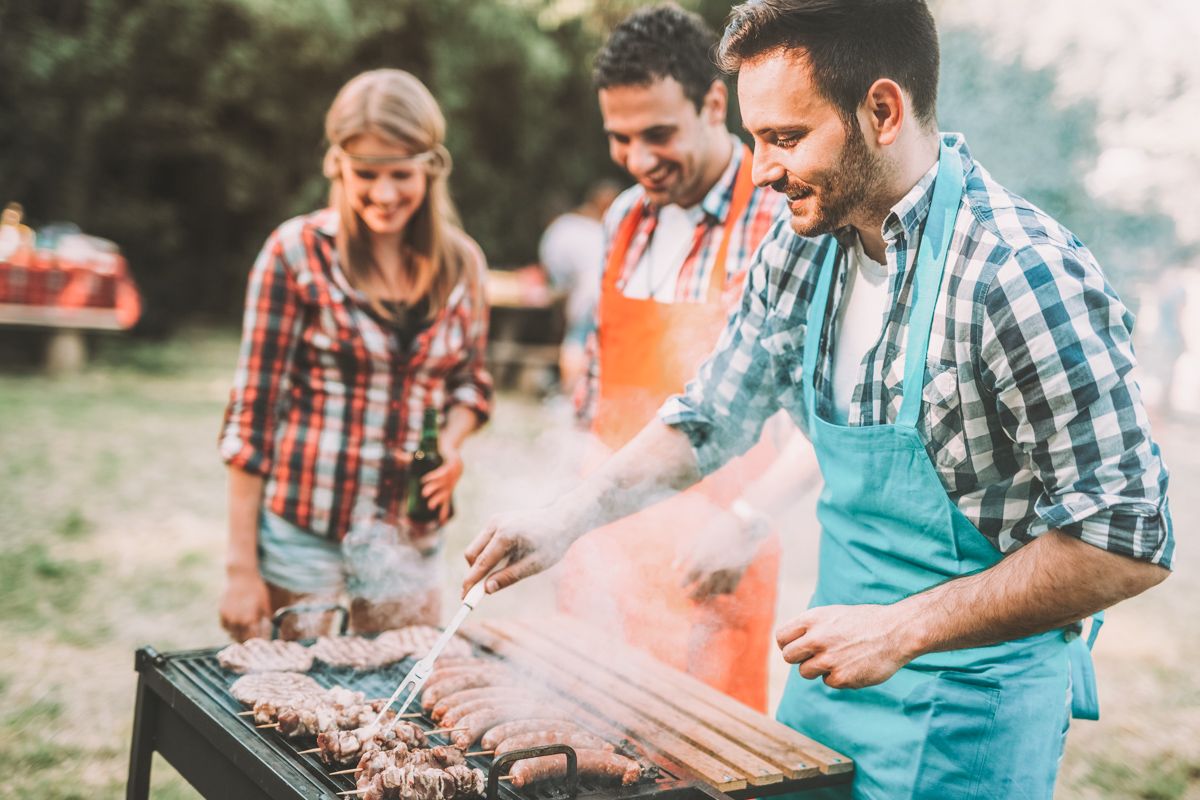 Two men and a woman, all wearing flannel shirts, stand around a grill with sausages and burgers sizzling on it.