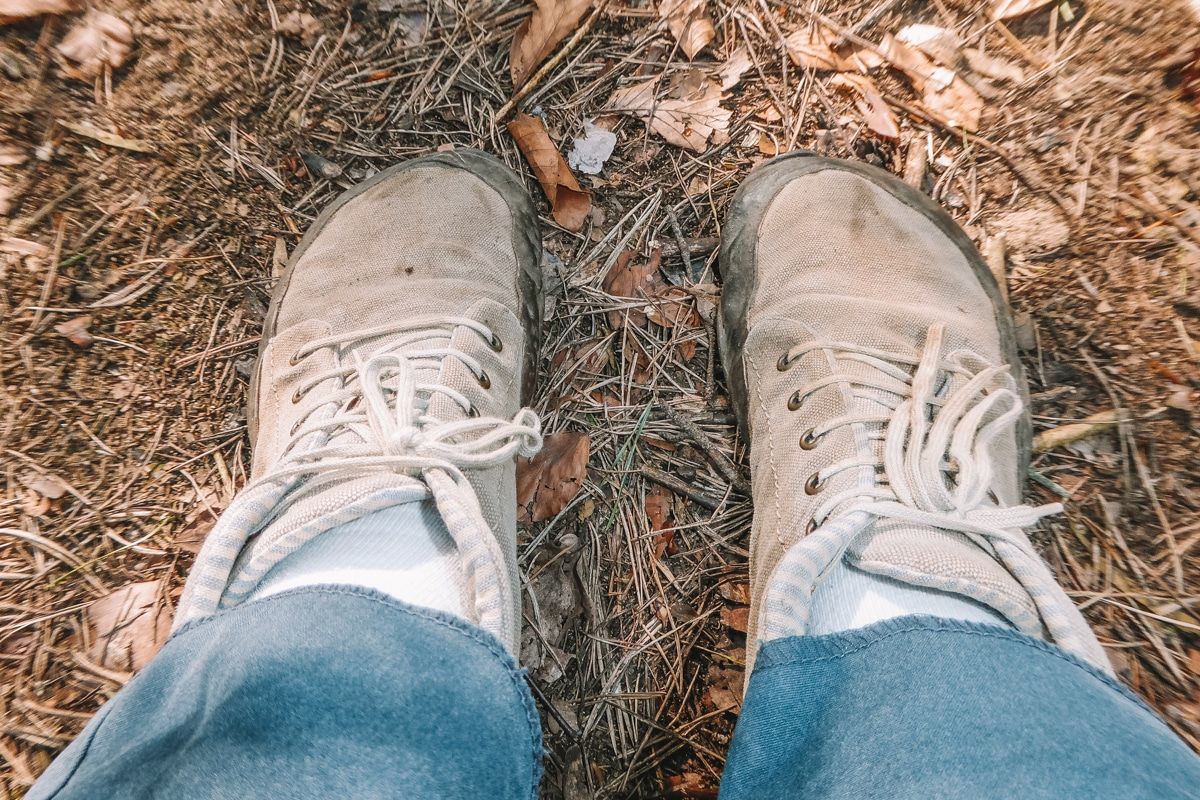 A pair of feet wearing light grey barefoot boots on a ground strewn with dry leaves and pine needles.