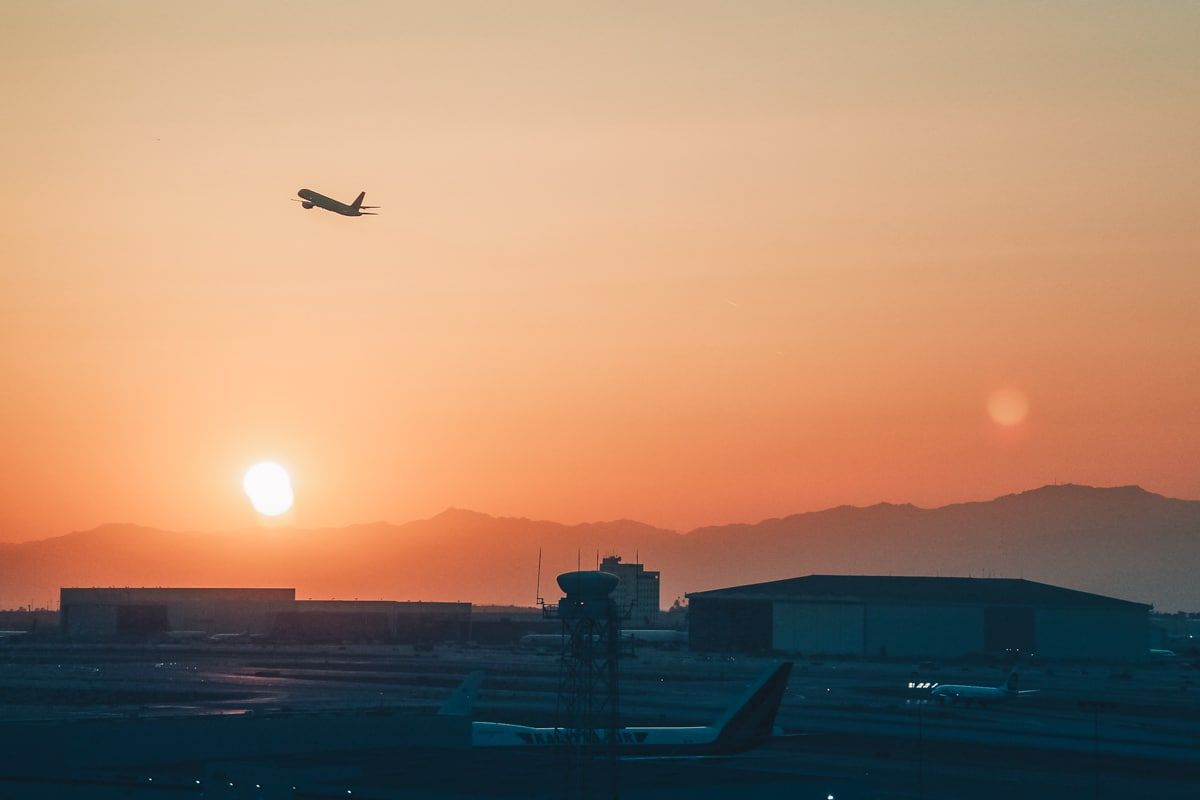 The silhouette of an airplane lifting off over the tarmac at Los Angeles International Airport (LAX) against an orange sunset.