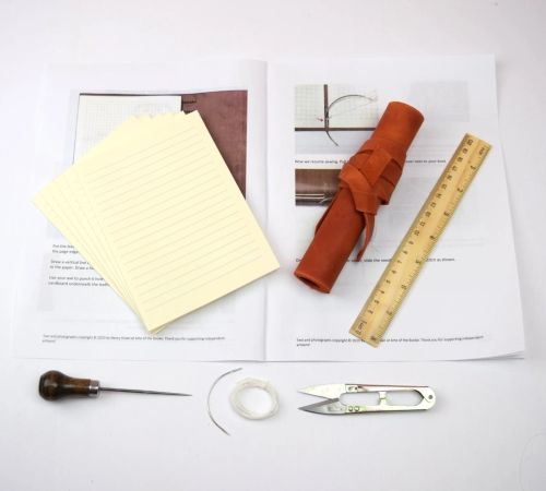 Materials for book binding laid out on a table, including leather, a ruler, line pages, scissors, string, an awl, and paper instructions.