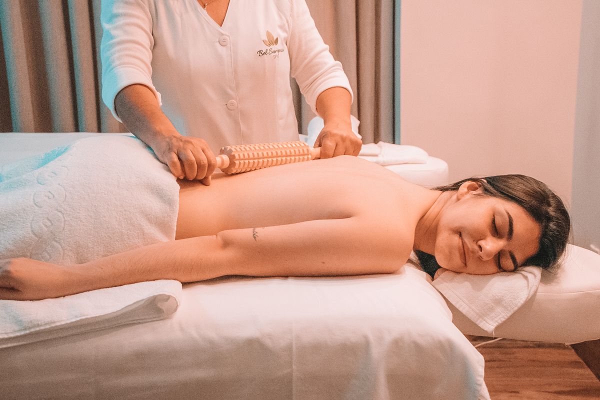 A woman lies on a white sheet on a a massage table and receives a massage with a textured wooden roller.