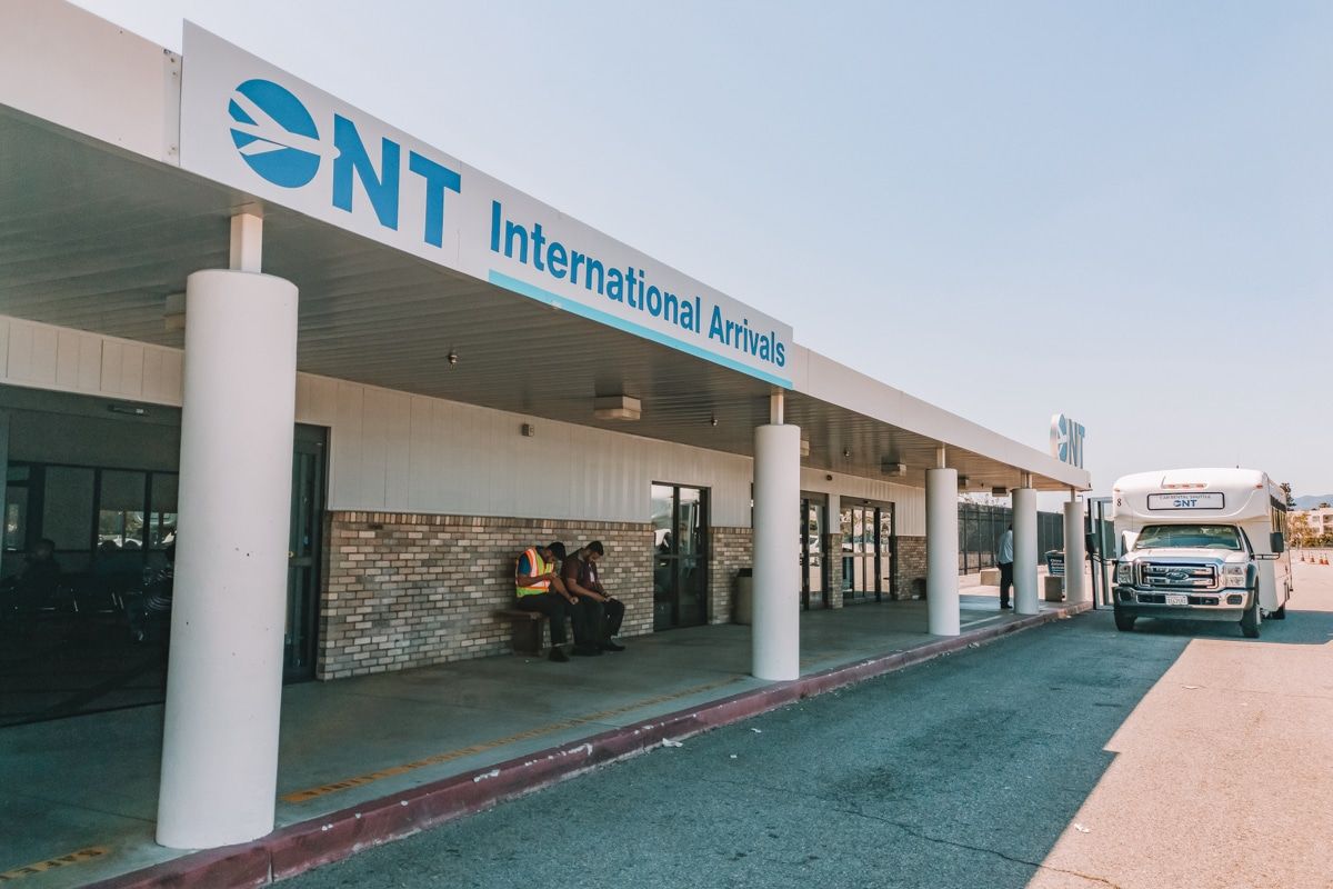 A small shuttle bus stopped outside of Ontario International Airport (ONT), while two workers sit on a bench in the shade.