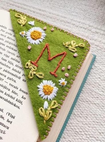 The corner of a book page enveloped in green fabric with daisies and the red letter "M" stiched.