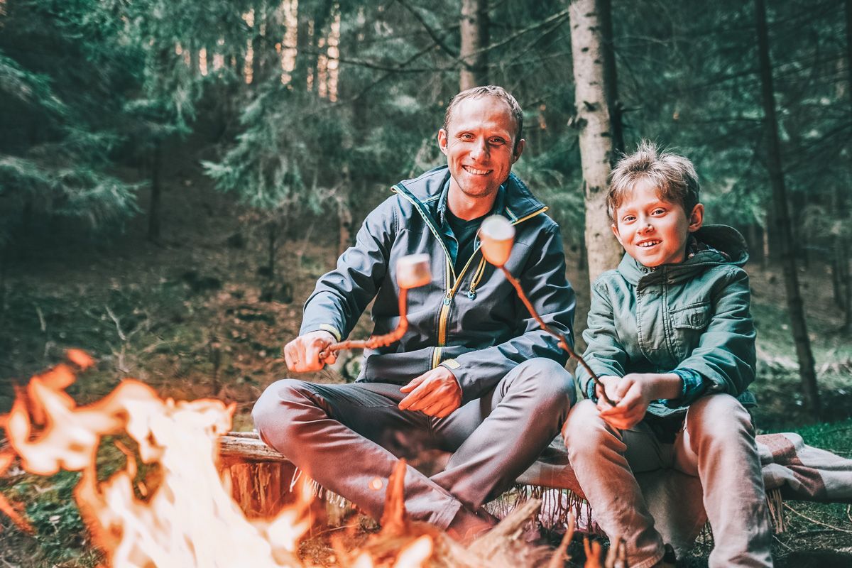 A smiling man and young boy wearing windbreakers roast marshmallows over a campfire with a forest in the background.