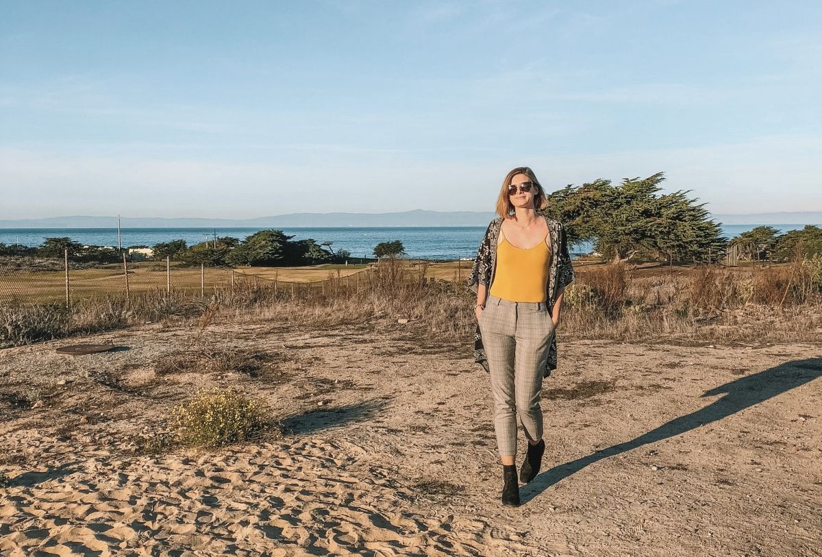A woman in tan plaid pants and a yellow shirt stands in a sandy landscape with the ocean visible in the distance.