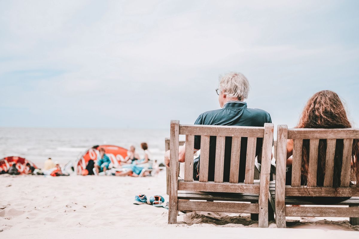 An older couple, seen from behind, sits on wooden beach chairs, looking out at a soft-focus ocean.