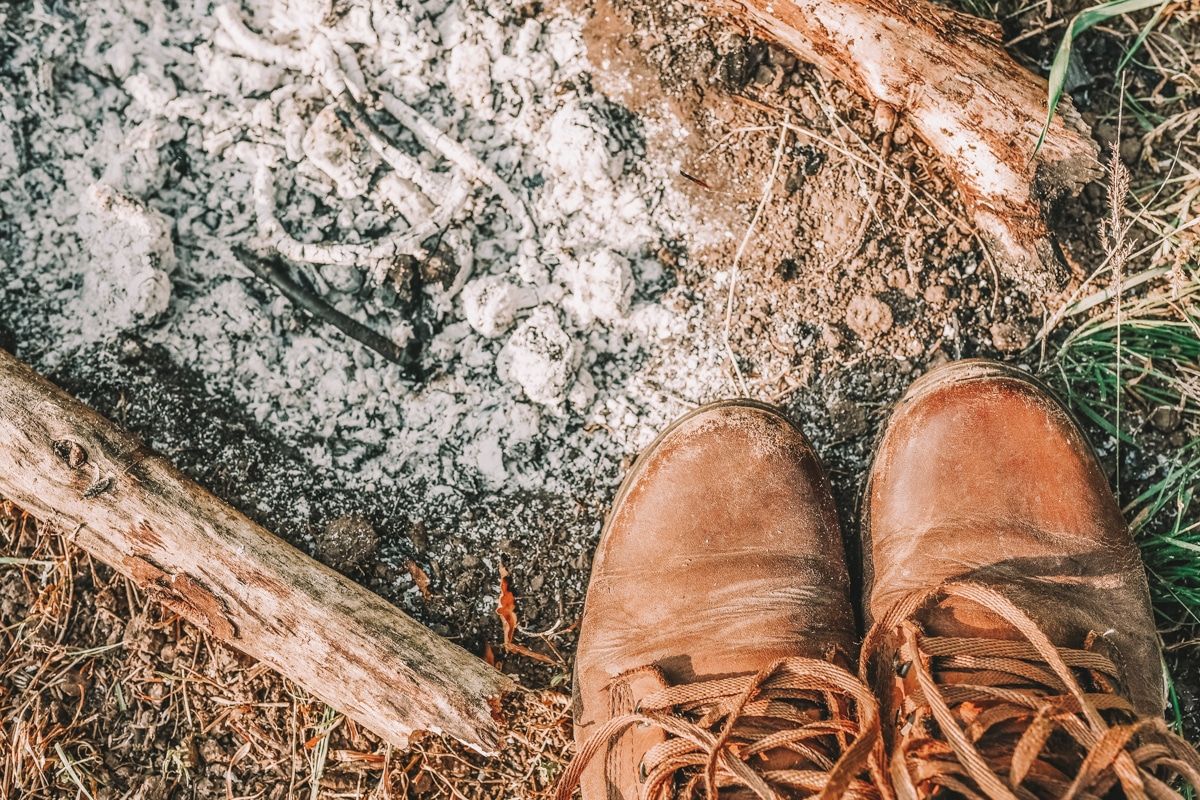 The toes of a pair of brown leather barefoot boots at the edge of a burned out campfire, full of ashes.