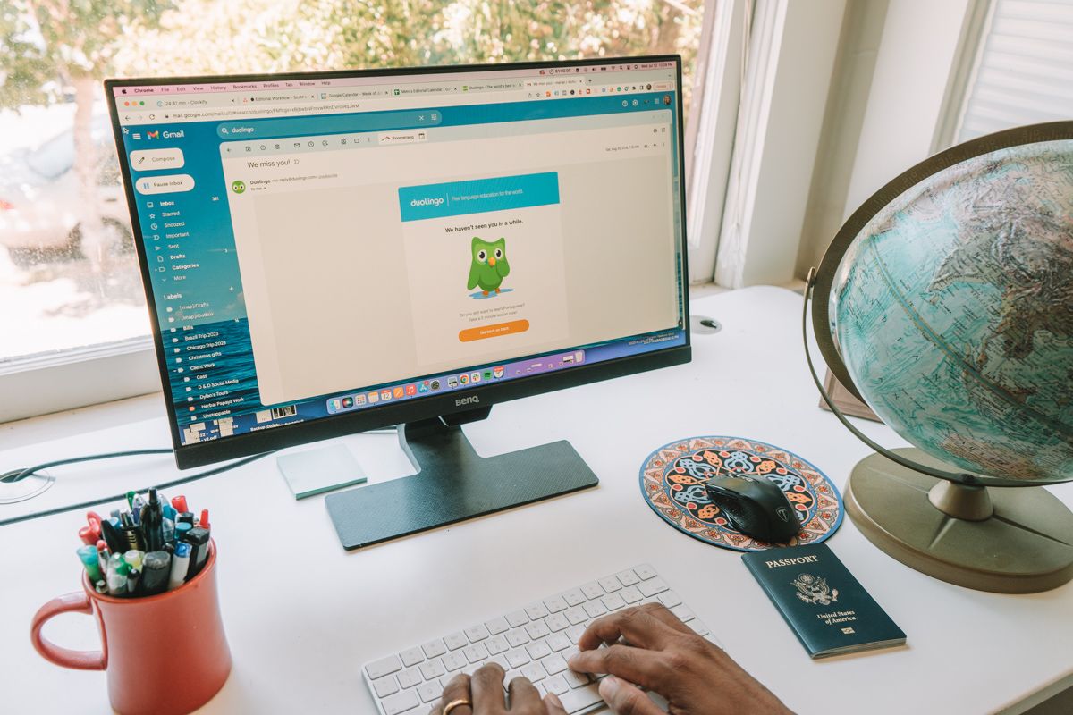 A white desk with the Duolingo website visible on the computer screen and a passport and globe sitting on the desk to right.