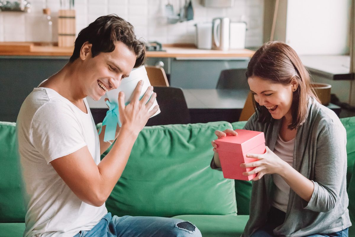 A brown-haired man and a brown-haired woman sit on a green couch facing each other, each excitedly preparing to open a gift.