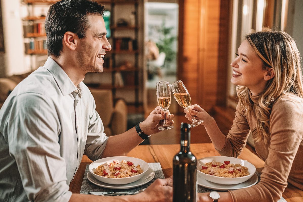 A brown-haired man clinks champagne glasses with a blonde woman in a beige shirt over plates of pasta, with a warmly-lit, soft-focus apartment interior in the background.