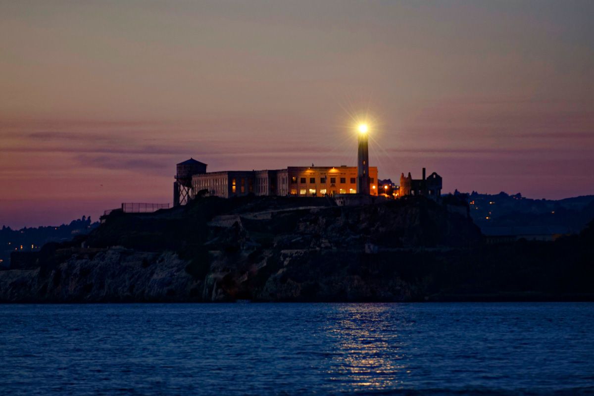 A view of Alcatraz at sunset illuminated orange against a pink sky, with the deep blue water in the foreground.