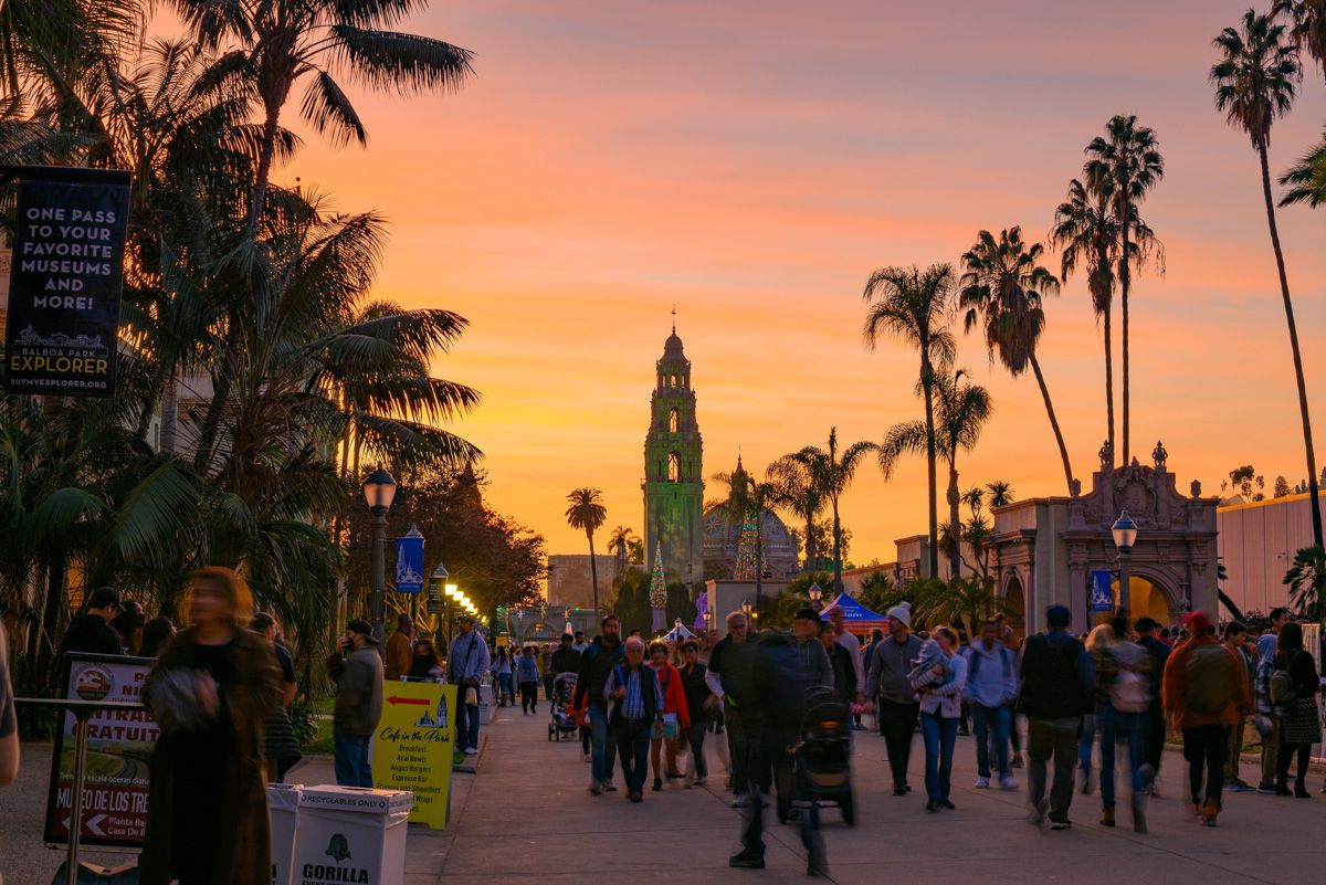A crowd of people strolling the palm-lined walkway leading to Balboa Park in San Diego beneath an orange and pink sunset.
