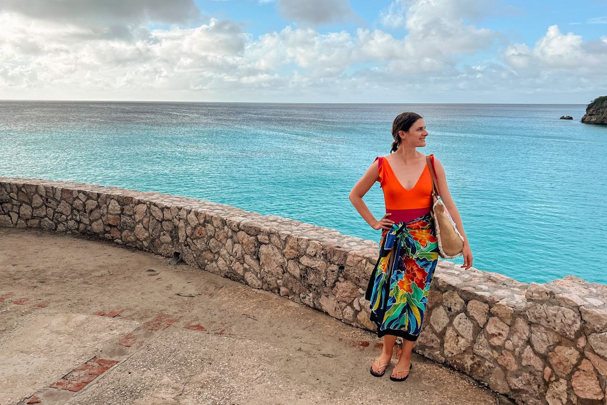 A woman wearing a bright orange top with a floral-printed sarong skirt stands with one hand on her hip next to a low stone wall overlooking a tropical, turquoise sea.