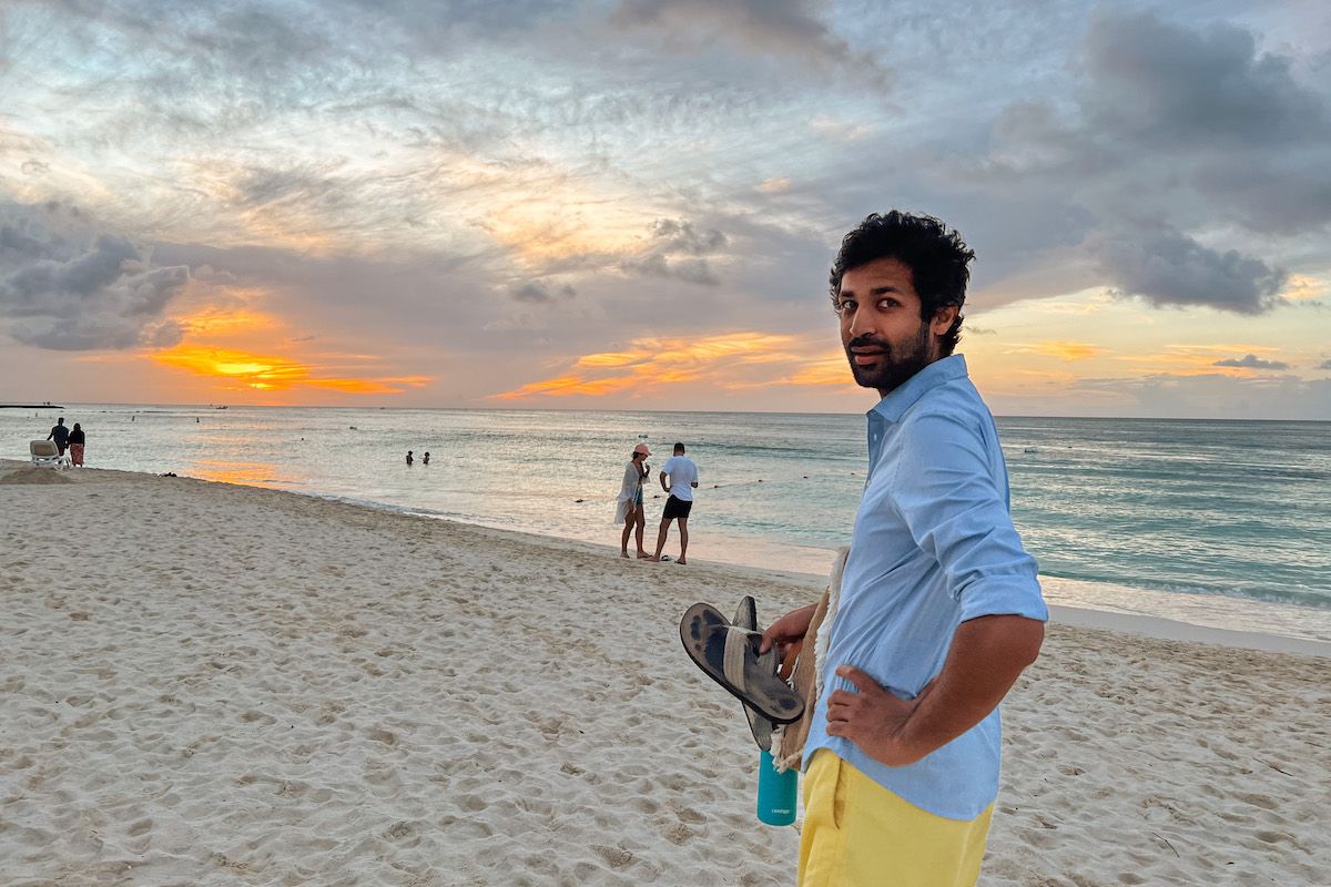 A man in a blue button-down and yellow shorts looks back over his shoulder while holding a pair of flip-flops on the beach with an orange and grey sunset behind him. 