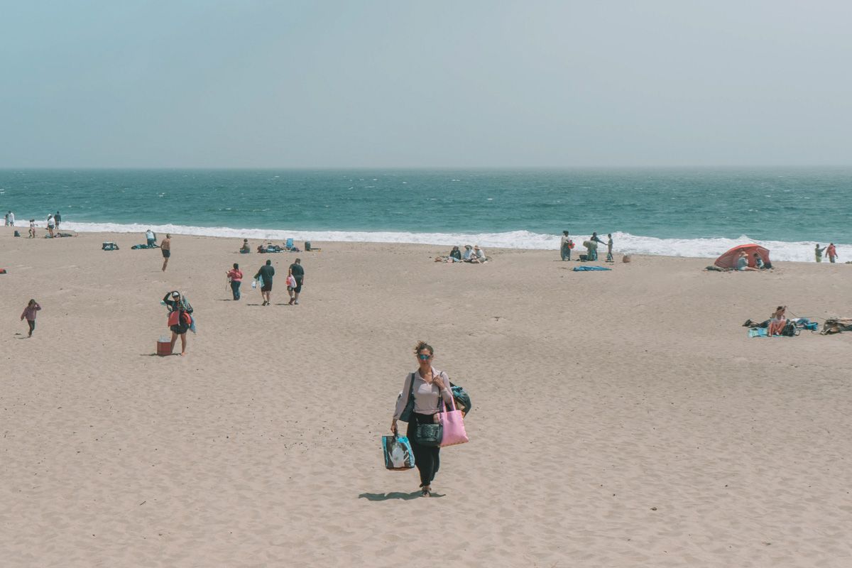A woman seen from a distance, walking on a sparsely-populated beach, carrying five tote bags.
