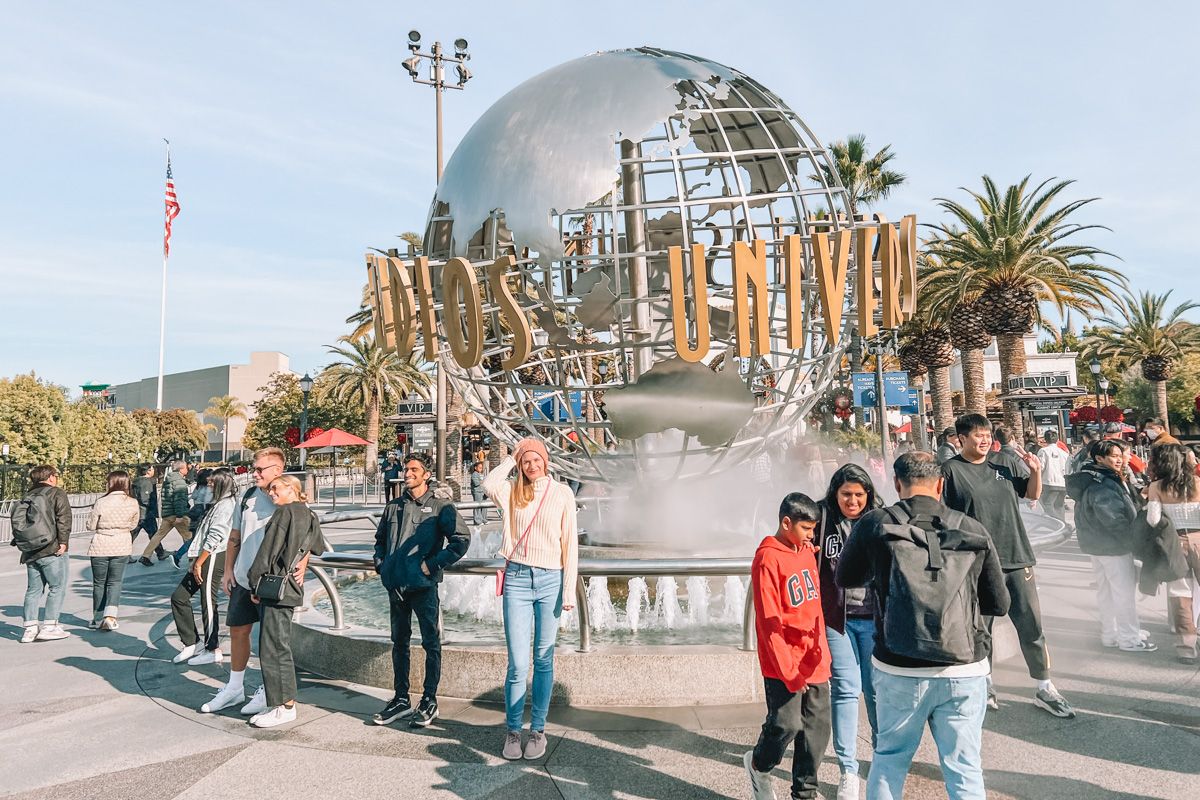 A young woman wearing blue jeans and a white sweater stands amidst a crowd in front of the Universal Studios globe statue on a clear day.
